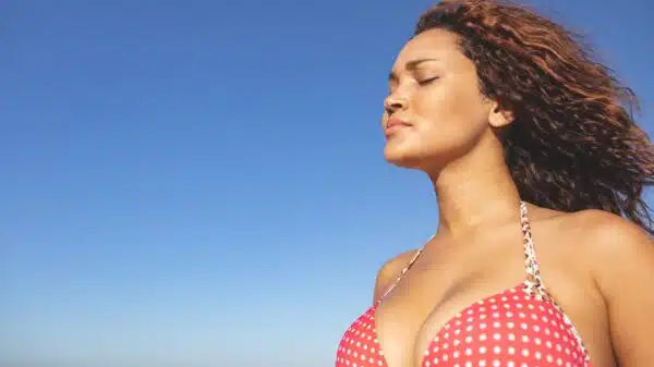 Side view of beautiful mixed race woman in bikini standing with eyes closed on the beach